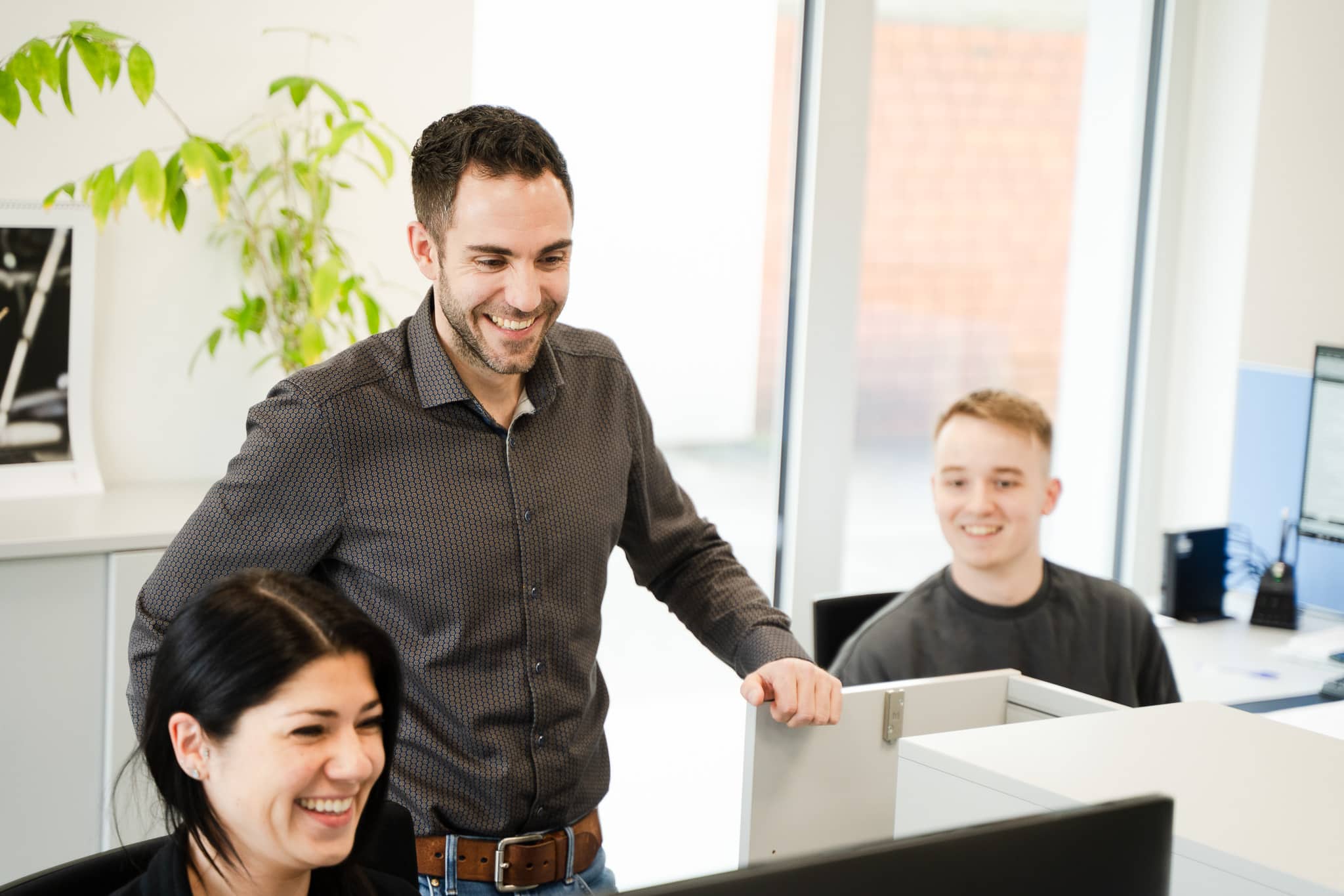 Drei lachende Mitarbeiter in einem modernen Büro arbeiten gemeinsam an einem Computerbildschirm.